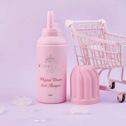 Pink bottle of 'Whipped Cream Lash Shampoo' Strawberry Scented, with a small pink shopping cart on a light purple background. Velvet Lash Co Formula 